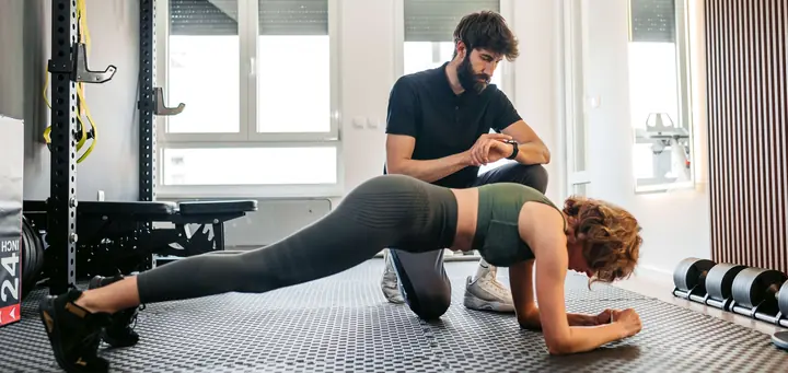 Trener prati vreme dok klijentkinja drži plank na podu studija